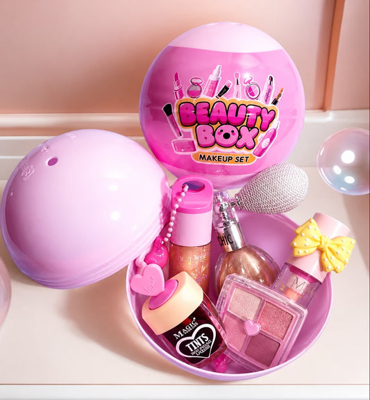 A pink beauty box makeup set displayed open on a light surface, with the matching round lid leaning beside it. The set includes a glittery pink container, a sparkly brush, a small yellow bow, a heart-shaped item, a square eyeshadow palette, and other pink cosmetic-style accessories.